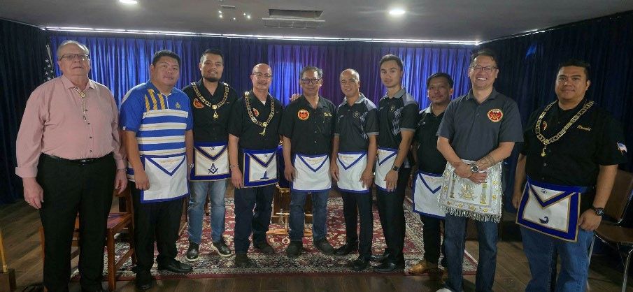 Ten lodge members in aprons and collars stand together for a group photo in front of blue curtains in the lodge hall.